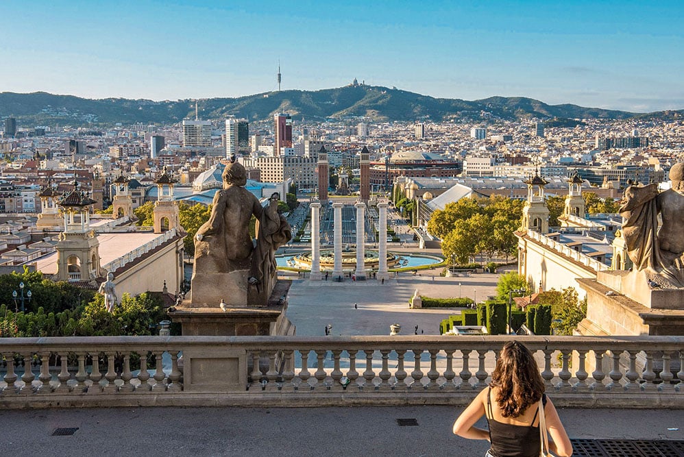 Ein Panoramablick über Barcelona aufgenommen vom Dach des Museu National d'Art de Catalunya. Auf dem Balkon ist eine Person zu sehen welche sich die Aussicht ansieht.