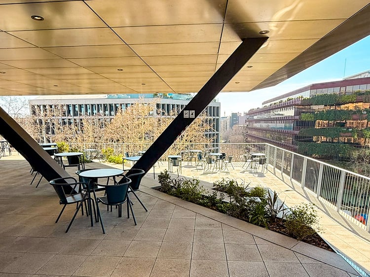 Eine weitläufige Dachterrasse der LCI Barcelona mit Blick auf die Stadtlandschaft des Viertels. Moderne Sitzgruppen aus schwarzen Metallstühlen und Tischen stehen unter einem massiven, schrägen Vordach, das von dunklen Stahlträgern gestützt wird.