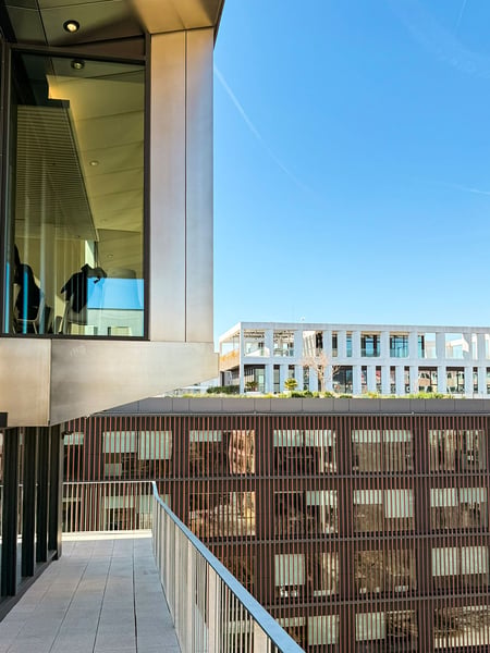Blick von einem Balkon der LCI Barcelona mit Blick auf die umliegende Architektur des Viertels. Links ragt ein Gebäudeteil mit einer metallischen Fassade und einem großen Fenster ins Bild, während im Hintergrund ein Gebäude mit einer braunen Lamellenstruktur unter einem blauen Himmel zu sehen sind.
