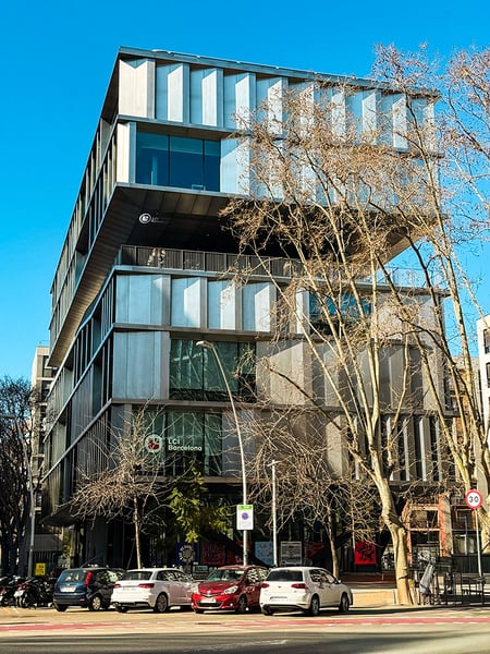 Frontale Ansicht der LCI Barcelona mit Fokus auf das markante, auskragende Obergeschoss. Die metallische Fassade reflektiert das Sonnenlicht, während im Vordergrund Autos an der Straße parken und hohe, blattlose Bäume einen Teil der Glasfront überschneiden.
