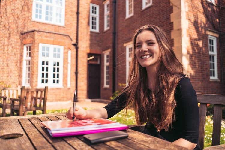 Ein Foto einer lächelnden Person mit Heften vor einem Backsteingebäude in England