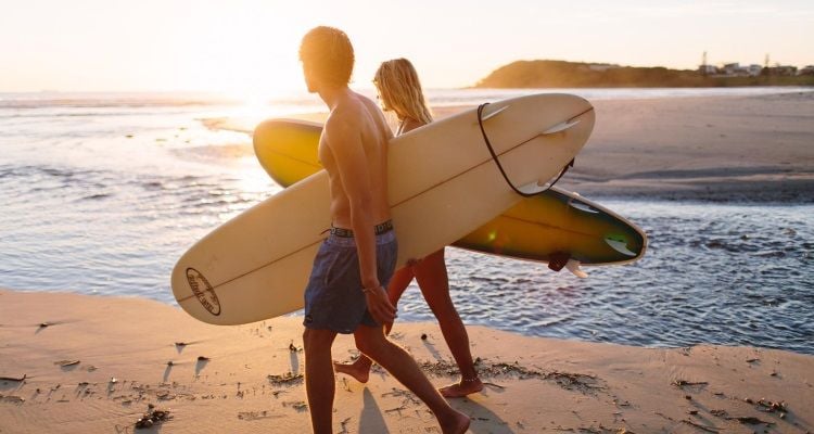 Zwei Leute haben Surfboards dabei und laufen einen Strand entlang