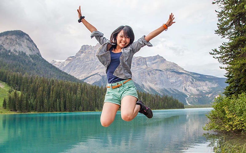 Frau springt am Emerald Lake: AIFS Kanada Adventure Trips inmitten atemberaubender Berg- und Seenlandschaft.