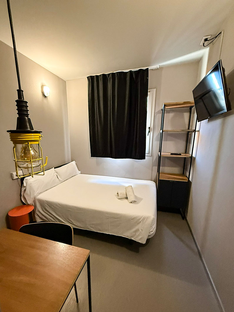 Ein gemütliches Einzelzimmer mit einem weiß bezogenen Bett, auf dem frische Handtücher bereitliegen. Zur Ausstattung gehören ein schwarzes Wandregal, ein an der Wand montierter Fernseher und ein kleiner Arbeitsplatz mit einer gelben Industrie-Hängelampe im Vordergrund.
