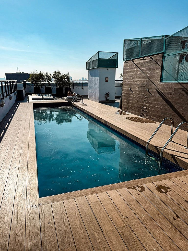 Ein schmaler, rechteckiger Dachpool mit klarem blauem Wasser, eingelassen in ein helles Holzdeck. Im Hintergrund laden Sonnenliegen zum Entspannen unter blauem Himmel ein, während moderne Sichtschutzelemente und eine braune Holzwand die Terrasse begrenzen.